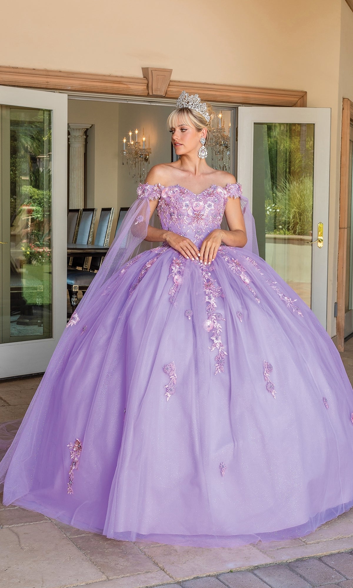 A woman in the Quinceanera Dress 1714 By Dancing Queen, a pastel purple ball gown with floral details and a tiara, stands in front of open double doors.